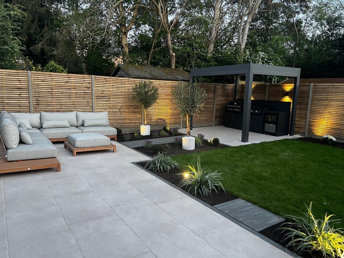 An RB Landscapes garden at dusk: porcelain terrace, lit olive trees, pergola kitchen and fresh lawn
