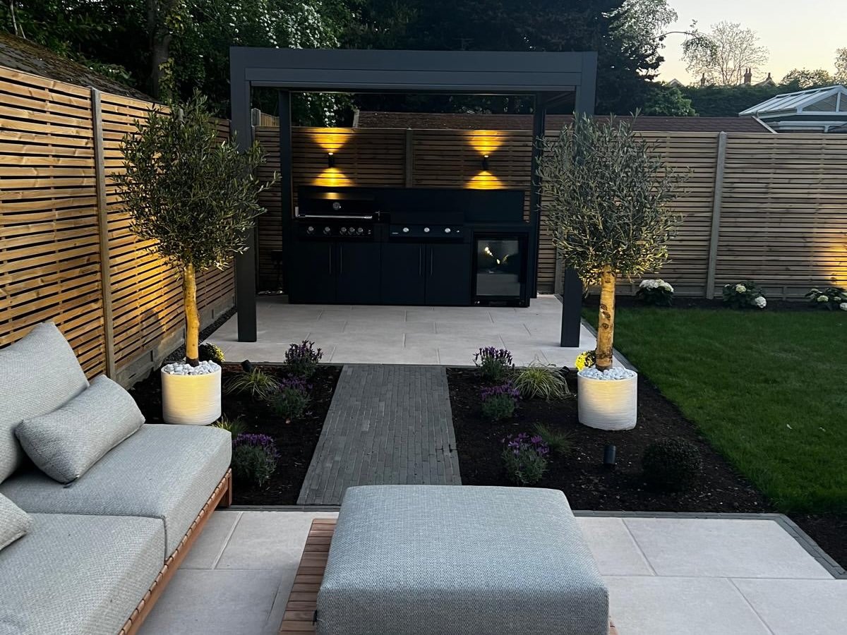 An outdoor kitchen under a pergola, lit by warm uplights at dusk